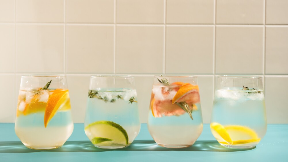 Summer refreshing sparkling drink with ice, fresh fruits and herbs: rosemary, thyme, orange, lime, lemon, grapefruit in transparent glasses on a blue background.