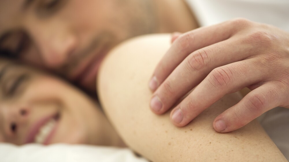 Couple cuddling together in bed, husbands hand on wifes shoulder, close-up